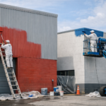 Workers painting industrial building exteriors with a ladder and lift